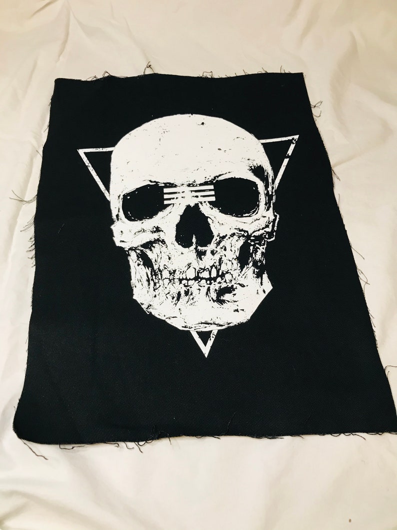 Op de afbeelding: Een zwarte stoffen patch met een wit schedelontwerp. De schedel heeft een driehoekige vorm eromheen en drie lijnen over de ogen.