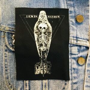 May include: Black and white embroidered patch featuring a skeleton inside a bomb. The bomb is surrounded by a triangle with the words "Lewis Mayhem" at the top.