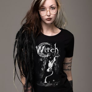May include: A black t-shirt with a white graphic of a crescent moon and the word "Witch" in a gothic font. The graphic also features a stylized figure with a long, flowing tail.
