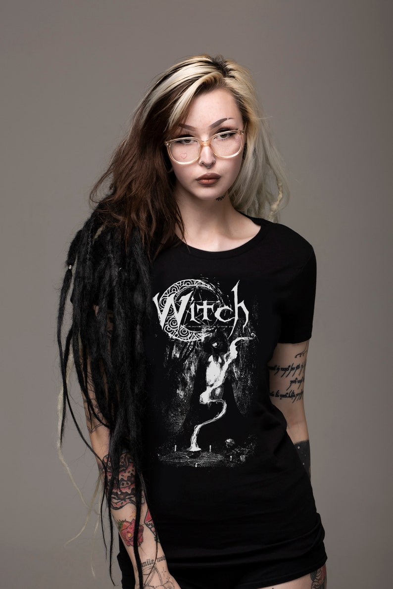 May include: A black t-shirt with a white graphic of a crescent moon and the word "Witch" in a gothic font. The graphic also features a stylized figure with a long, flowing tail.