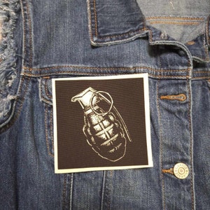 May include: A white and black hand grenade patch on a brown background. The patch is sewn onto a denim jacket.