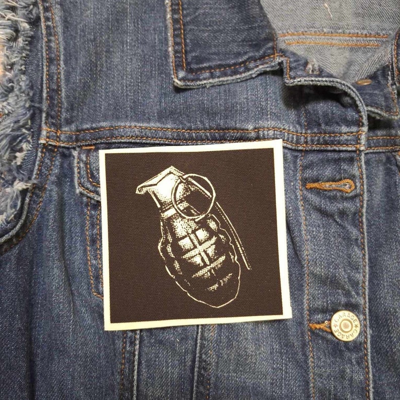 May include: A white and black hand grenade patch on a brown background. The patch is sewn onto a denim jacket.