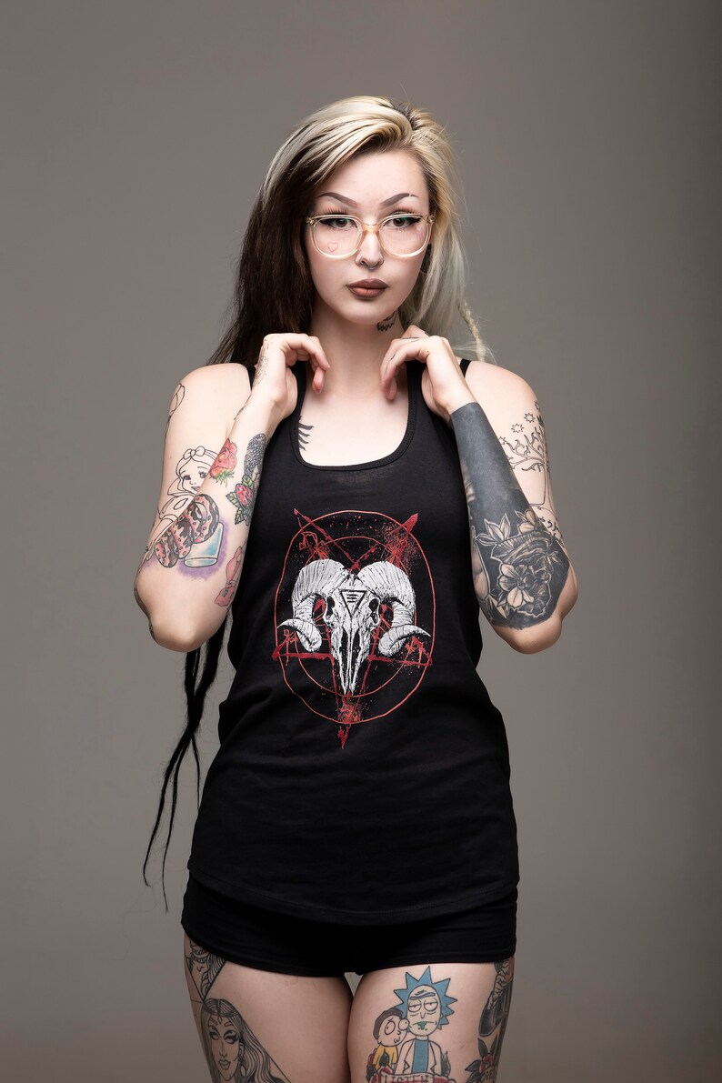 May include: A woman with black and blonde hair wearing a black tank top with a red and white graphic of a skull with horns and a pentagram. She has several tattoos on her arms and legs.