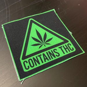 May include: Square black patch with a green triangle containing a cannabis leaf. The text "CONTAINS THC" is printed in white. The patch is made of fabric.