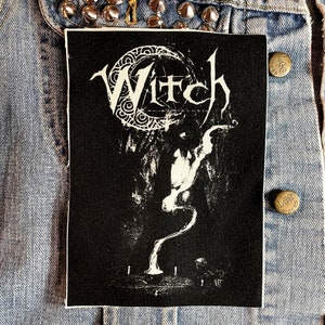 May include: Black and white iron-on patch with the word "Witch" in a stylized font. The patch features a crescent moon with a swirling design and a stylized hand with smoke rising from it.
