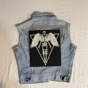 K&ouml;nnte beinhalten: Eine hellblaue Jeansweste mit einem schwarzen Patch, der ein Skelett mit Fl&uuml;geln in einem Dreieck zeigt. Die Weste ist mit silbernen Nieten verziert.