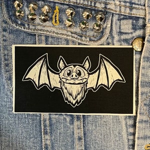 Può includere: Toppa termoadesiva in bianco e nero con un pipistrello dei cartoni animati con un'espressione amichevole e le ali spiegate.