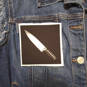 May include: A black and white iron-on patch with a white knife on a black background.