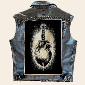 Può includere: Una toppa in bianco e nero con un'immagine stilizzata di un cuore trafitto da un pugnale. Il cuore è circondato da un effetto di luce radiante. La toppa è cucita su un giubbotto di jeans.