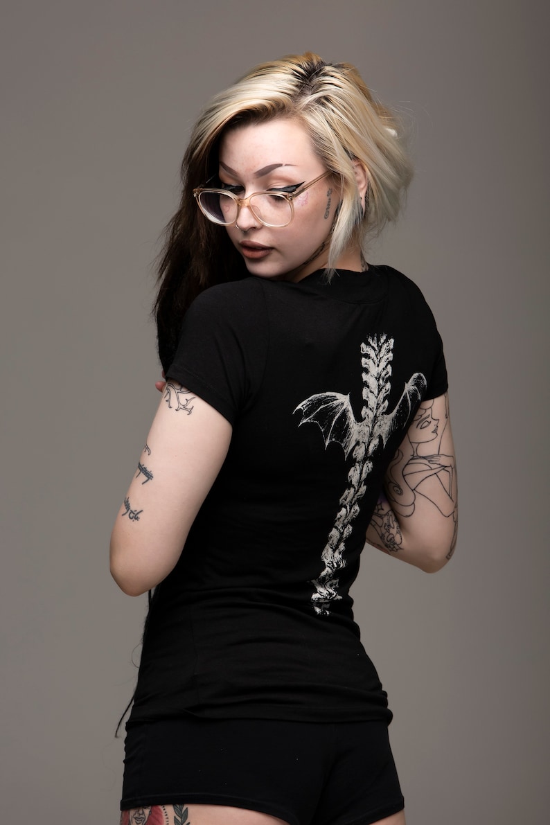 K&ouml;nnte beinhalten: Eine Frau mit blonden und schwarzen Haaren tr&auml;gt ein schwarzes T-Shirt mit einem wei&szlig;en Skelett und Fl&uuml;geln auf dem R&uuml;cken. Sie hat mehrere Tattoos auf ihren Armen und Beinen.