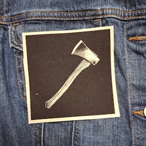 May include: A black and white patch with a white axe design on a black background. The axe is angled diagonally from the bottom left to the top right of the patch.