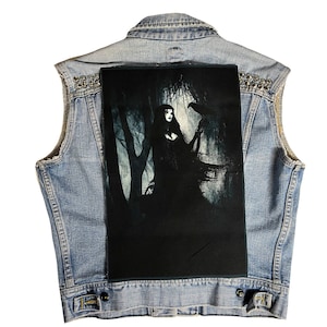 May include: A denim vest with a black patch featuring a woman in a dark forest with a raven. The vest has silver studs around the collar and shoulders.