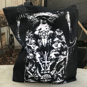 May include: Black tote bag with a white graphic print of a skull with a pentagram and the number 666. The skull is surrounded by two figures with wings.