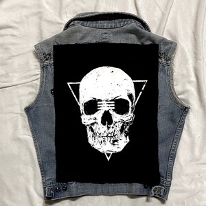 Op de afbeelding: Een zwart denim vest met een witte schedel en driehoek patch. De schedel heeft een verweerde look en draagt een zonnebril.