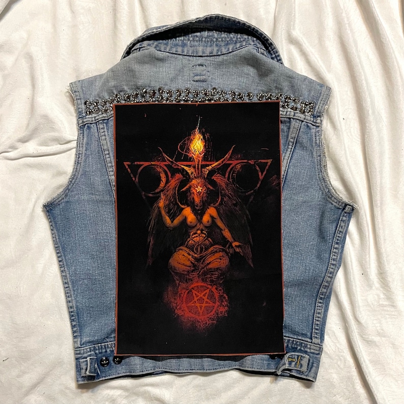 K&ouml;nnte beinhalten: Hellblaue Jeansweste mit einem gro&szlig;en, rechteckigen Aufn&auml;her mit einer detaillierten Illustration von Baphomet. Die Weste ist mit silberfarbenen Nieten am Kragen verziert. Das Baphomet-Bild ist in Rot-, Orange- und Schwarzt&ouml;nen gehalten.