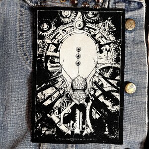 May include: Black and white patch featuring a detailed illustration of a skull with gears and machinery. The text "LEWIS MAYHEM" is at the bottom of the patch.