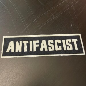 May include: A rectangular patch with the word "ANTIFASCIST" in bold, white letters. The text is centered on a black background, framed by a thin white border. The patch is likely made of fabric.