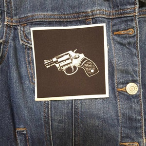 Può includere: Un'immagine in bianco e nero di una pistola a tamburo su una toppa marrone scuro cucita su una giacca di jeans blu.
