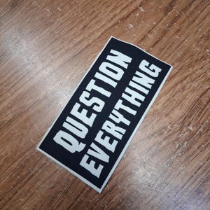 Può includere: Una toppa rettangolare nera con la scritta "QUESTION EVERYTHING" in grassetto bianco maiuscolo. La toppa è angolata su una superficie di legno con un motivo a venature naturali.