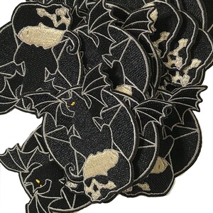 Pode incluir: Um monte de patches de ferro bordados em preto e branco com uma caveira com asas de morcego. A caveira &eacute; delineada em branco e tem um olho branco. As asas de morcego tamb&eacute;m s&atilde;o delineadas em branco.