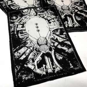 May include: Black and white embroidered patch featuring a detailed illustration of a creature with a skull-like face surrounded by gears and machinery. The patch has a textured fabric surface.