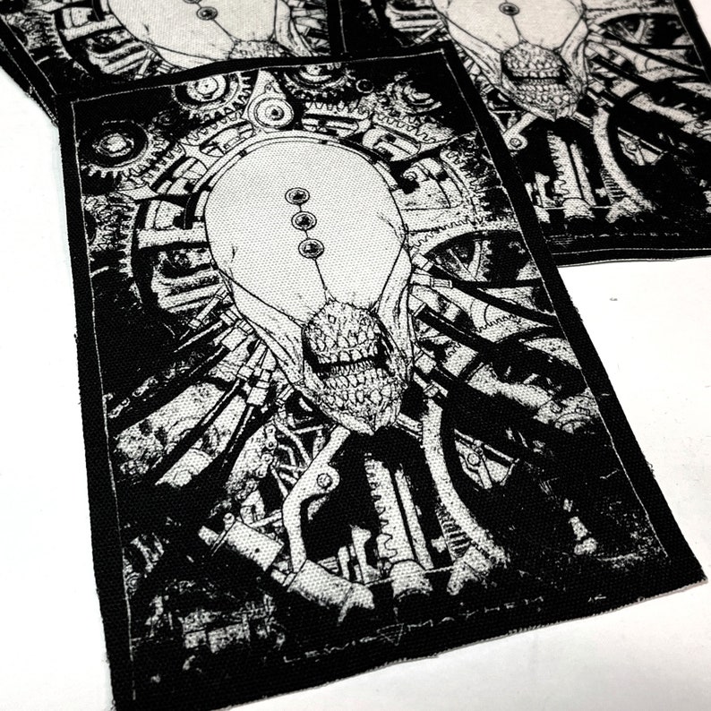 May include: Black and white embroidered patch featuring a detailed illustration of a creature with a skull-like face surrounded by gears and machinery. The patch has a textured fabric surface.