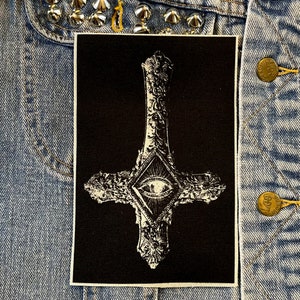 May include: Black and white embroidered patch of a pentagram with an eye in the center. The pentagram is surrounded by ornate details.