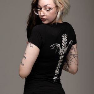 K&ouml;nnte beinhalten: Eine Frau mit blonden und schwarzen Haaren tr&auml;gt ein schwarzes T-Shirt mit einem wei&szlig;en Skelett und Fl&uuml;geln auf dem R&uuml;cken. Sie hat mehrere Tattoos auf ihren Armen und Beinen.