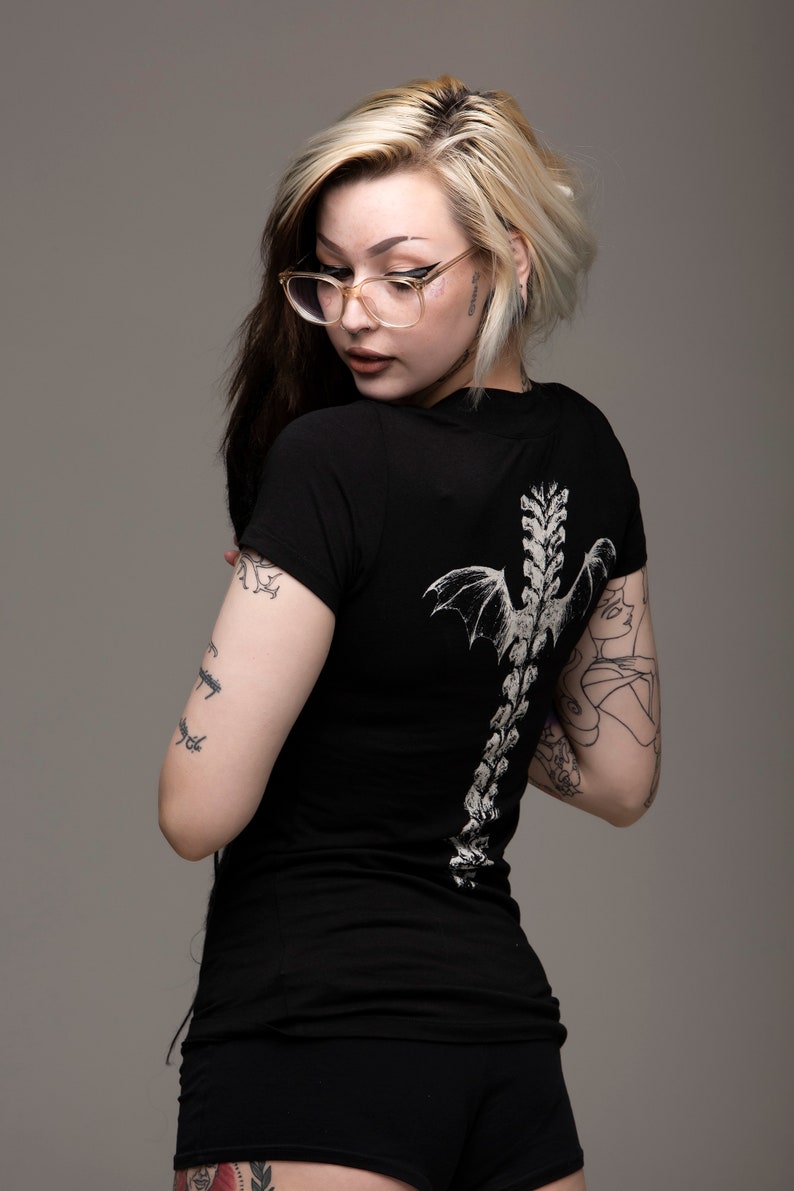 K&ouml;nnte beinhalten: Eine Frau mit blonden und schwarzen Haaren tr&auml;gt ein schwarzes T-Shirt mit einem wei&szlig;en Skelett und Fl&uuml;geln auf dem R&uuml;cken. Sie hat mehrere Tattoos auf ihren Armen und Beinen.