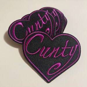 May include: Black heart-shaped embroidered patches with the word "Cunty" in a cursive, magenta font. The patches have a magenta border and are stacked on top of each other.
