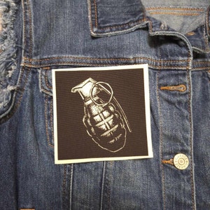 May include: A black and white embroidered patch of a grenade with a pin pulled. The patch is sewn onto a denim jacket.
