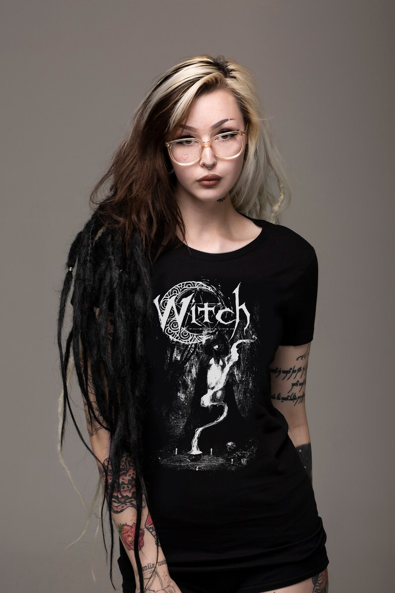 May include: A black t-shirt with a white graphic of a crescent moon and the word "Witch" in a gothic font. The graphic also features a stylized figure with a long, flowing tail.
