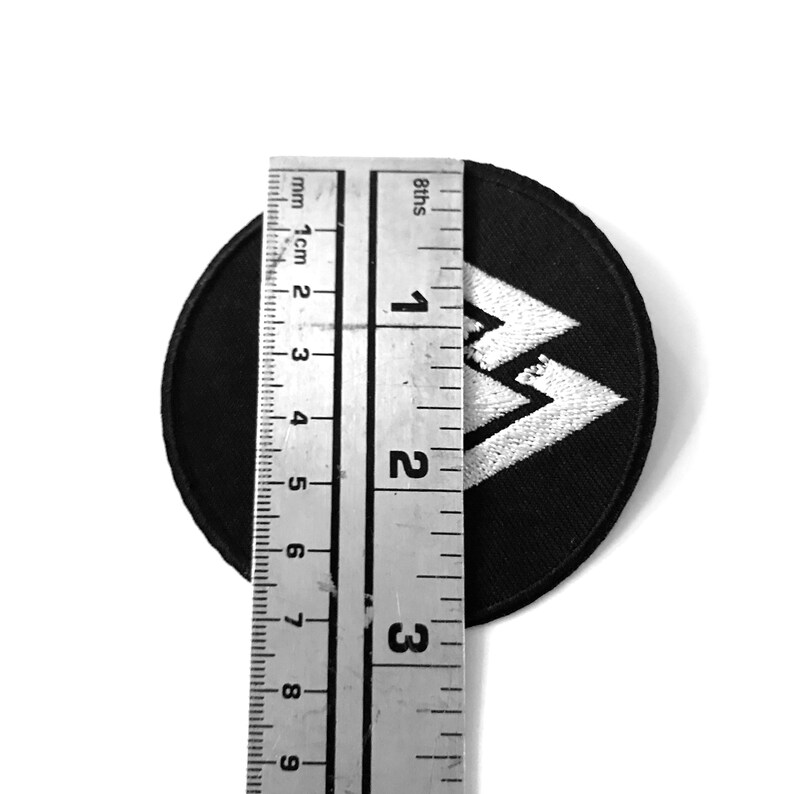 May include: A black and white embroidered patch with a stylised mountain design. The patch is approximately 5 cm in diameter.