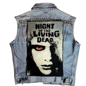 Könnte beinhalten: Eine hellblaue Jeansweste mit einem Grafikmotiv von "Night of the Living Dead". Die Weste hat eine Reihe silberner Nieten am oberen Rand und einen Knopfverschluss. Die Grafik ist ein Schwarz-Weiß-Bild eines Zombies mit roten Augen.