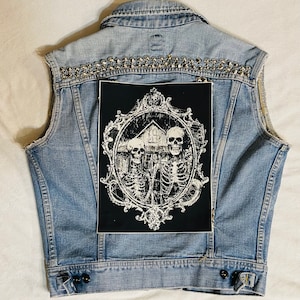 May include: A blue denim vest with silver studs and a black patch featuring a white ornate frame with three skeletons and a house inside.