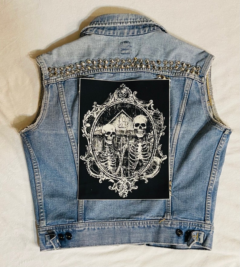 May include: A blue denim vest with silver studs and a black patch featuring a white ornate frame with three skeletons and a house inside.