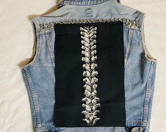 Op de afbeelding: Een lichtblauw spijkergilet met een grote rechthoekige zwarte patch op de achterkant met een witte grafische print van een menselijke ruggengraat. Het gilet is voorzien van een rij zilverkleurige metalen studs langs het bovenste rugpand.