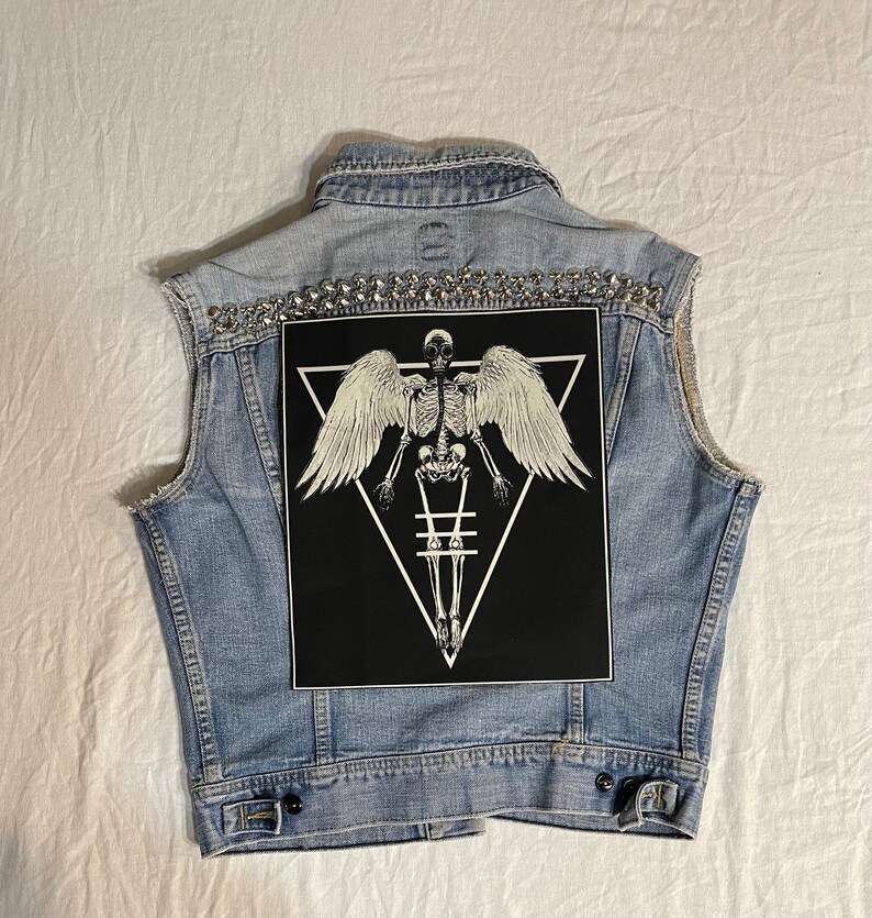 K&ouml;nnte beinhalten: Eine hellblaue Jeansweste mit silbernen Nieten und einem schwarzen Patch mit einem wei&szlig;en Skelett mit Fl&uuml;geln in einem Dreieck.