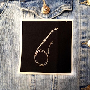 Può includere: Una toppa bianca e nera con un'immagine bianca di una frusta su uno sfondo nero.