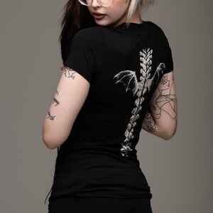 K&ouml;nnte beinhalten: Schwarzes, figurbetontes T-Shirt mit einem wei&szlig;en Aufdruck einer Wirbels&auml;ule mit Fl&uuml;geln auf dem R&uuml;cken.