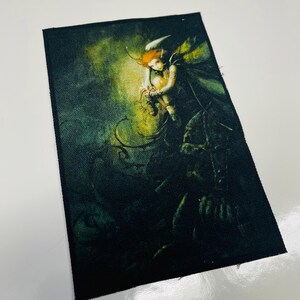 May include: A black fabric patch featuring a colourful image of a fairy with wings, holding a small creature. The fairy is surrounded by green vines and a glowing light.