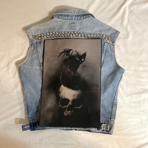 May include: A black cat with wings sits atop a skull, the image is printed on a black fabric patch. The patch is attached to a denim vest with silver studs.
