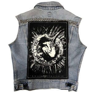 May include: A sleeveless denim jacket with a black and white graphic patch on the back. The patch features a detailed illustration of a figure within a circular design. The collar is embellished with silver studs. The text "I LEWIS MAYHEM" is visible.