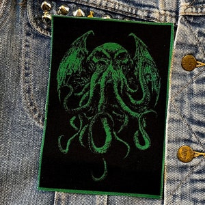 Puede incluir: Parche rectangular negro con un diseño verde de Cthulhu. El parche presenta una ilustración detallada de la criatura mítica con alas y tentáculos. El parche está bordeado con costuras verdes y se muestra en una chaqueta vaquera.