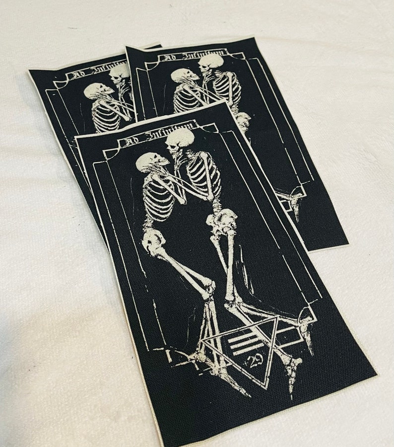 K&ouml;nnte beinhalten: Drei schwarze Stoffpatches mit wei&szlig;en gestickten Skeletten, die sich die H&auml;nde halten. Der Text "Ad Infinitum" befindet sich &uuml;ber den Skeletten. Der untere Patch hat ein Pentagramm mit der Zahl "23" darin.