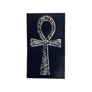 Puede incluir: Un parche de tela negro con un s&iacute;mbolo Ankh bordado en blanco.