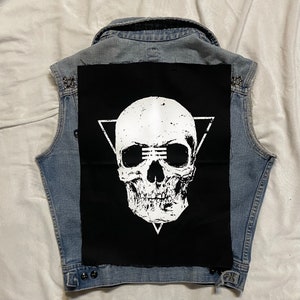 Op de afbeelding: Een zwart denim vest met een witte schedel patch op de rug. De schedel bevindt zich in een driehoek en heeft een verweerde look. Het vest heeft zilveren studs rond de kraag en zakken.