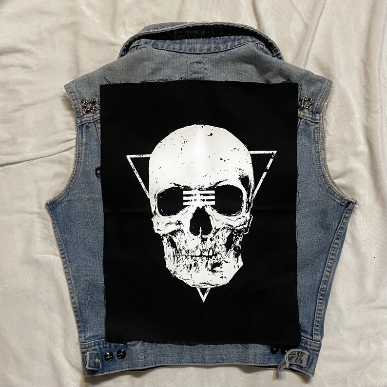Op de afbeelding: Een zwart denim vest met een witte schedel patch op de rug. De schedel bevindt zich in een driehoek en heeft een verweerde look. Het vest heeft zilveren studs rond de kraag en zakken.