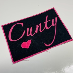 Könnte beinhalten: Ein schwarzer Stoff-Patch mit rosa Nähten an den Rändern. Der Patch hat das Wort "Cunty" in rosa kursiven Buchstaben mit einem rosa Herz unter dem Wort.
