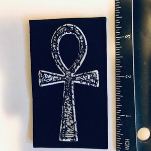 Puede incluir: Un parche de tela negro con un s&iacute;mbolo Ankh bordado en blanco. El parche mide aproximadamente 5 cm de ancho y 7,5 cm de alto.